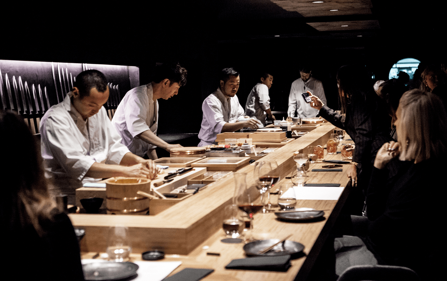 A group of chefs crafting sushi at a wooden bar with diners sitting watching.